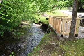 A wooden deck overlooking a stream with a stone pathway and a grassy area. - Creek Front Lodge • Private Setting • Fast Internet • Firepit (Hayesville)
