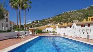 Piscina com água azul cristalina, rodeada por edifícios brancos com detalhes amarelos, palmeiras e um cenário de colinas.