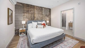 A bedroom with a brick accent wall, a bed with white bedding, a nightstand with a lamp, and a door leading to a bathroom. - The BlueJay Vintage Suite (No Window) (Grand Rapids)