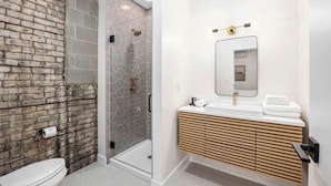 A modern bathroom with a brick wall, a glass shower enclosure, and a wooden vanity with a mirror. - The BlueJay Vintage Suite (No Window) (Grand Rapids)
