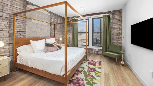 Ein modernes Schlafzimmer mit einem Himmelbett, einer Ziegelwand, einem großen Fenster mit Vorhängen und einem flachbildfernseher, der an der Wand montiert ist.