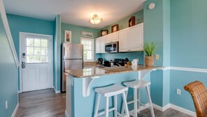 Une cuisine aux murs de couleur turquoise, avec des meubles blancs, un réfrigérateur en acier inoxydable, un four à micro-ondes et un comptoir en granit. On y trouve deux tabourets de bar blancs et une chaise en osier.