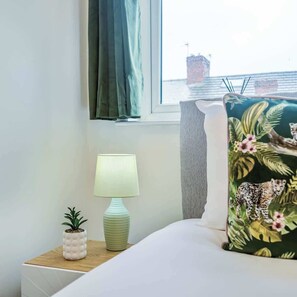 A neatly made bed with a floral pillow, a small table with a lamp and a potted plant, and a window with a view of a building across the water.