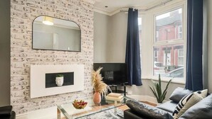 A living room with a brick wall, a mounted TV, a sofa, a coffee table, and a window with curtains. - Willow House - 2-Bedroom House In Merseyside (Merseyside)