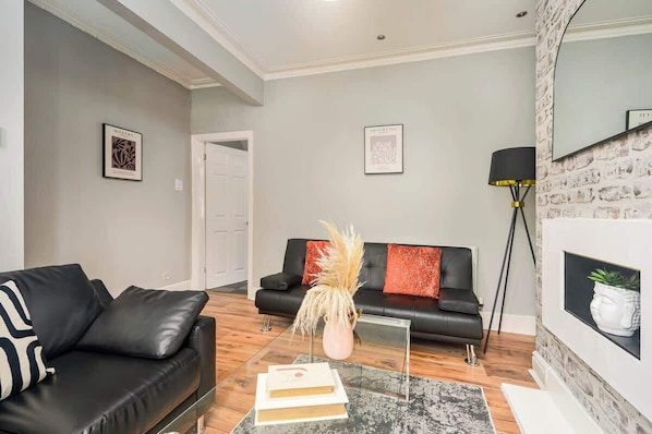 A modern living room with a black leather sofa, a glass coffee table, and a fireplace with a white mantel.