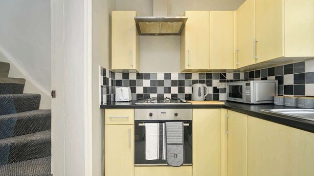 A compact kitchen with yellow cabinets, a black and white checkered backsplash, a stove, a microwave, and a toaster. - Willow House - 2-Bedroom House In Merseyside (Merseyside)