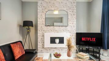 Una sala de estar con un sofá negro, una lámpara de pie, un televisor mostrando 'NETFLIX' y un espejo colgado en la pared.