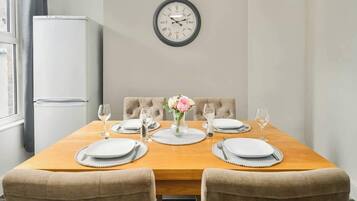 A dining room with a wooden table set for four, a clock on the wall, and a refrigerator in the background.