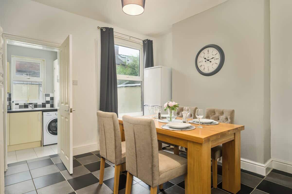 A dining area with a wooden table set for four, a clock on the wall, and a view of the outside through a window.