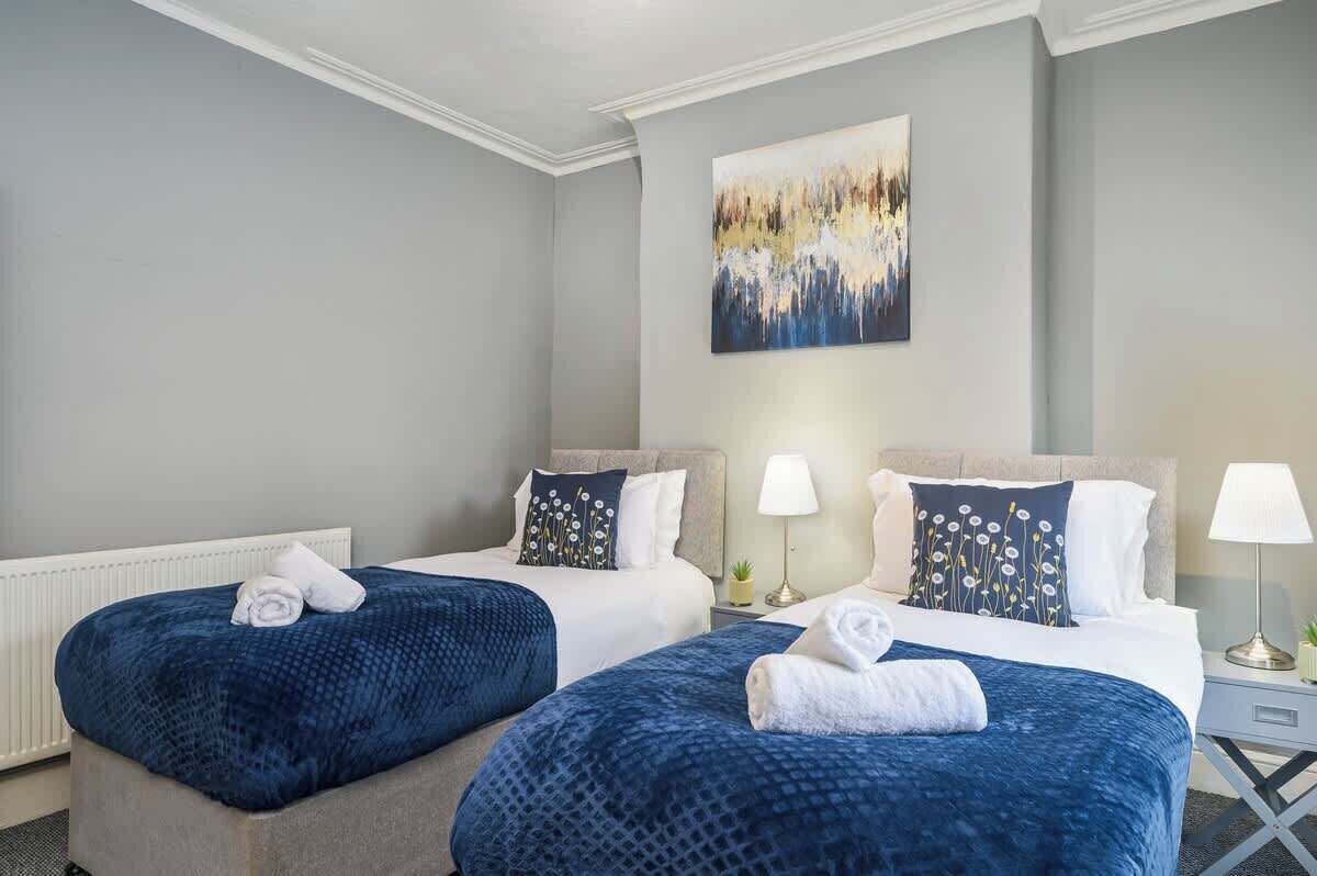 Habitación de hotel con dos camas, cada una con cubrecama azul y ropa de cama blanca. Hay un cuadro en la pared sobre las camas, y una mesita de noche con una lámpara entre ellas.
