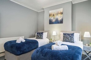 A hotel room with two beds, each with a blue bedspread and white linens. There is a painting on the wall above the beds, and a small table with a lamp between the beds. - Willow House - 2-Bedroom House In Merseyside (Merseyside)