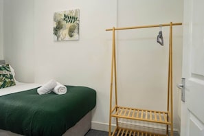 A room with a bed, a green blanket, a wooden drying rack, and a wall-mounted artwork.