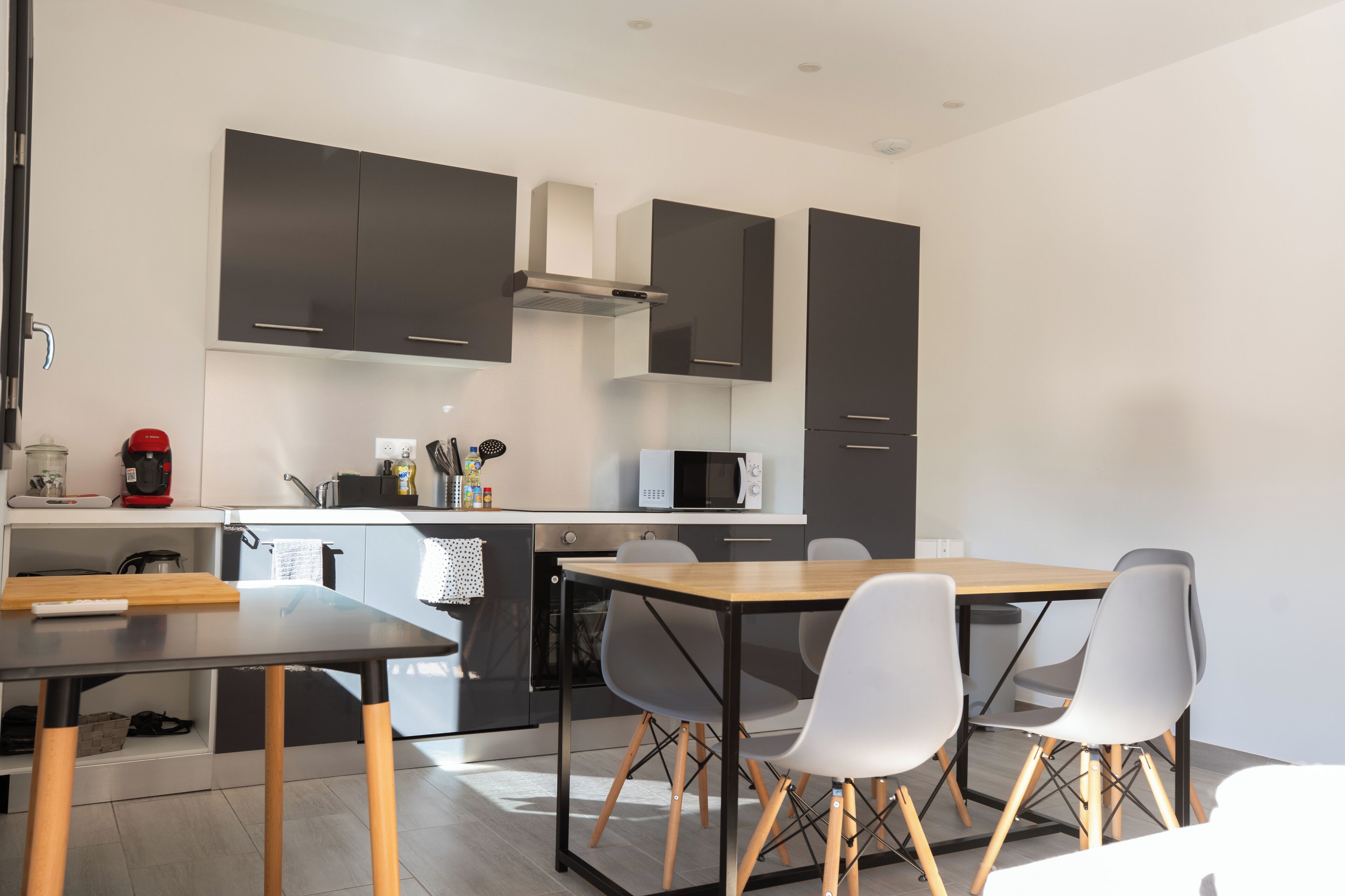 Een moderne keuken met donkere kastjes, een magnetron en een koffiezetapparaat. Er staat een houten tafel met vier stoelen.