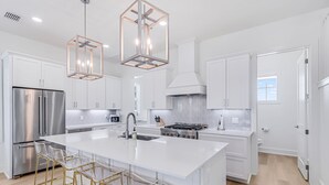 A modern kitchen with white cabinetry, a stainless steel refrigerator, a large island with bar stools, and pendant lighting. - 3-story home near the beach with outdoor pool, golf, deck, grill, streaming, W/D (Port Aransas)