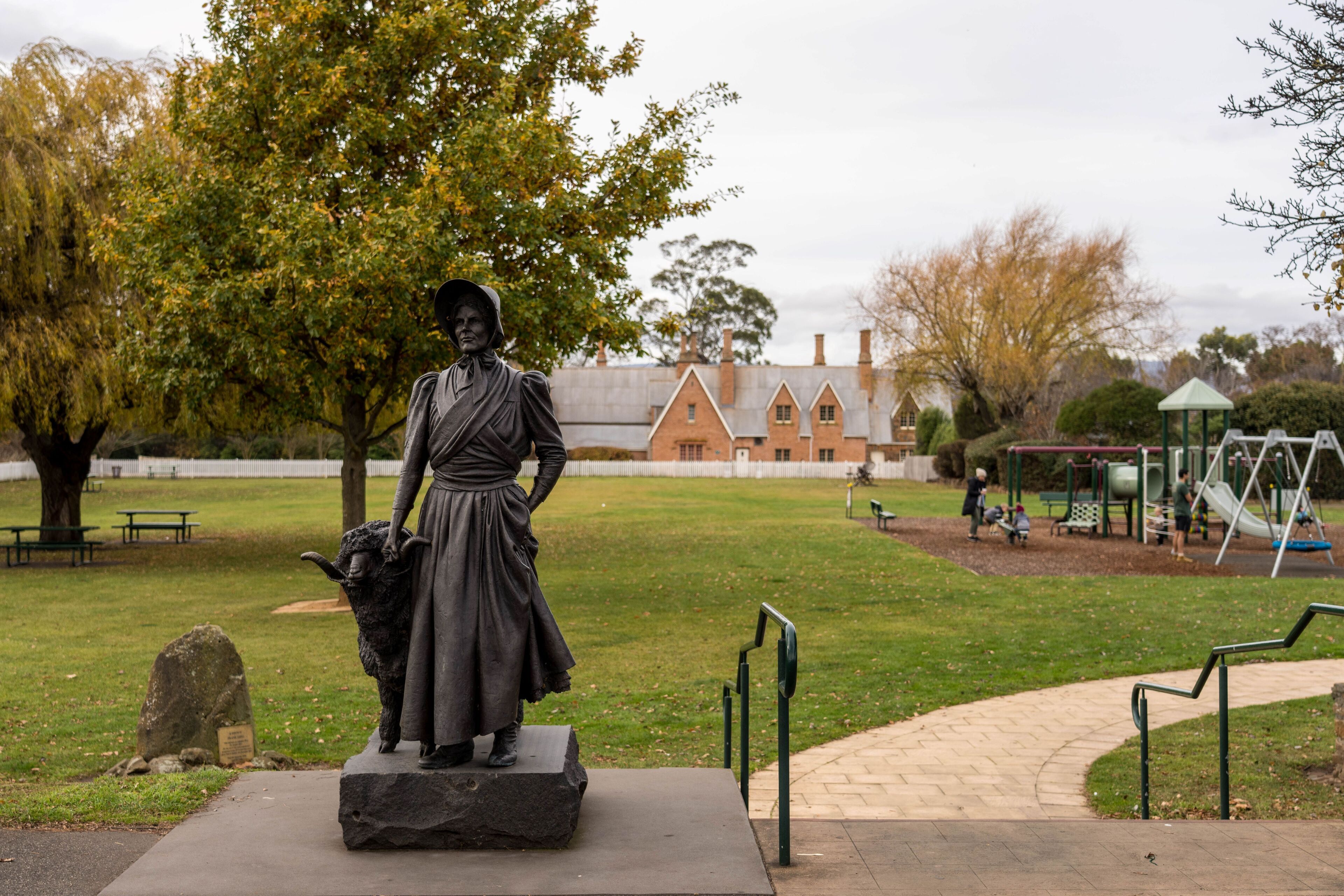 A statue of a woman holding a lamb in a park with a playground and a building in the background.