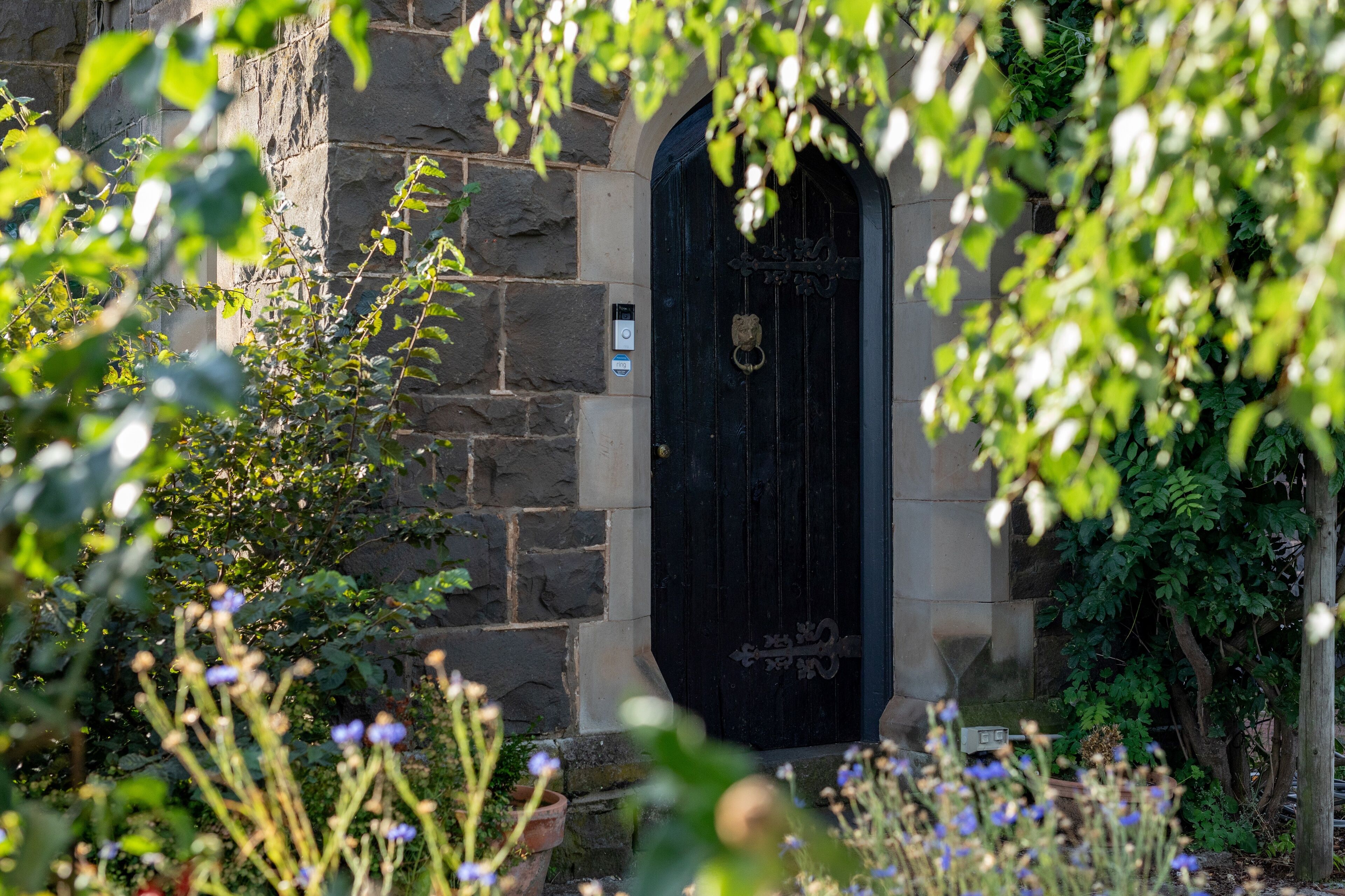 A stone building with a black door, surrounded by greenery and flowers.