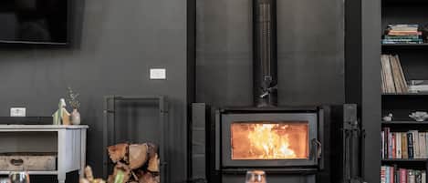 Ein gemütliches Wohnzimmer mit Kamin, Flachbildfernseher, Bücherregal und einem Esstisch mit Obstkorb und Weingläsern.