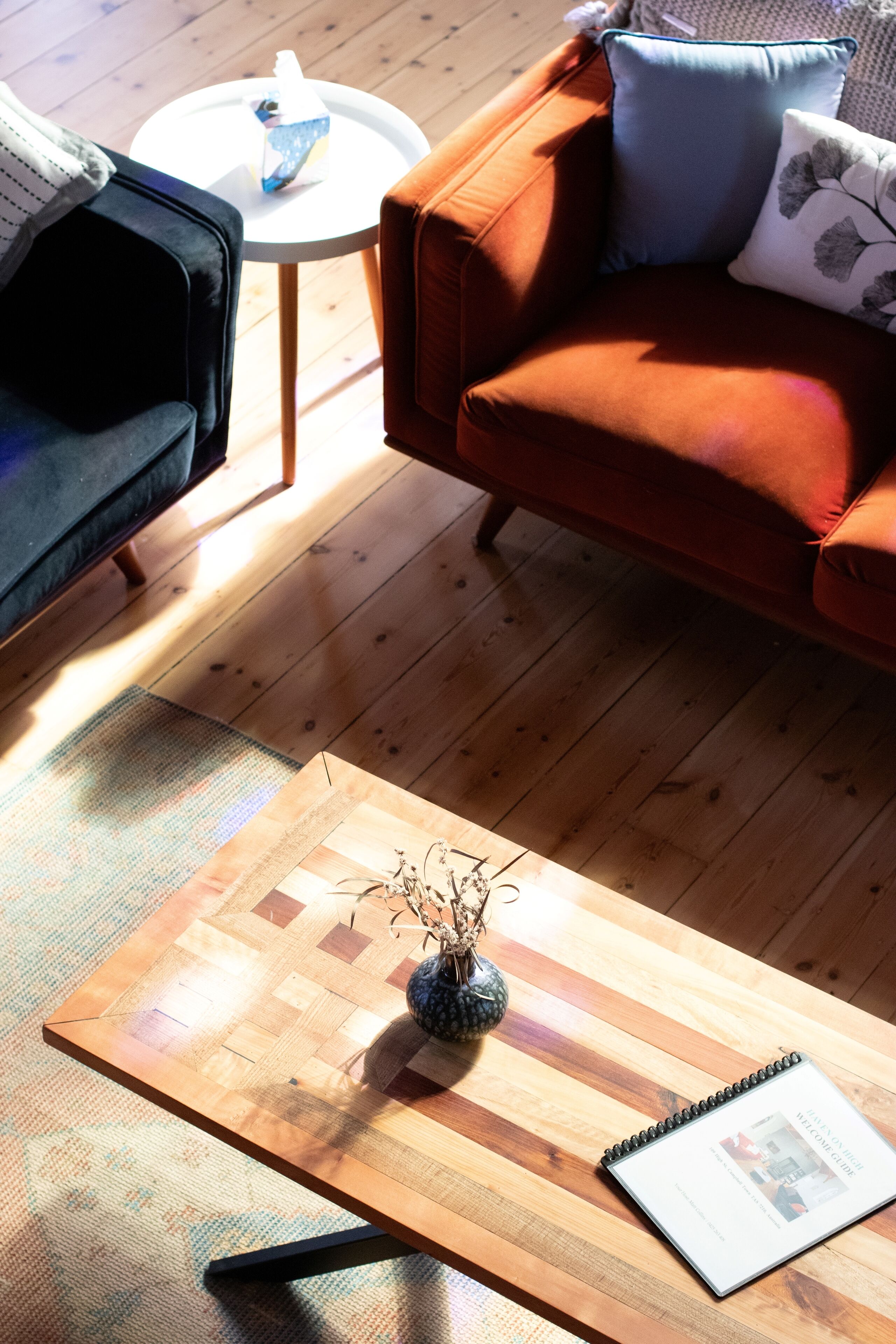 A wooden coffee table with a vase and a notebook, a red sofa with a pillow, and a small round table with a blue object.