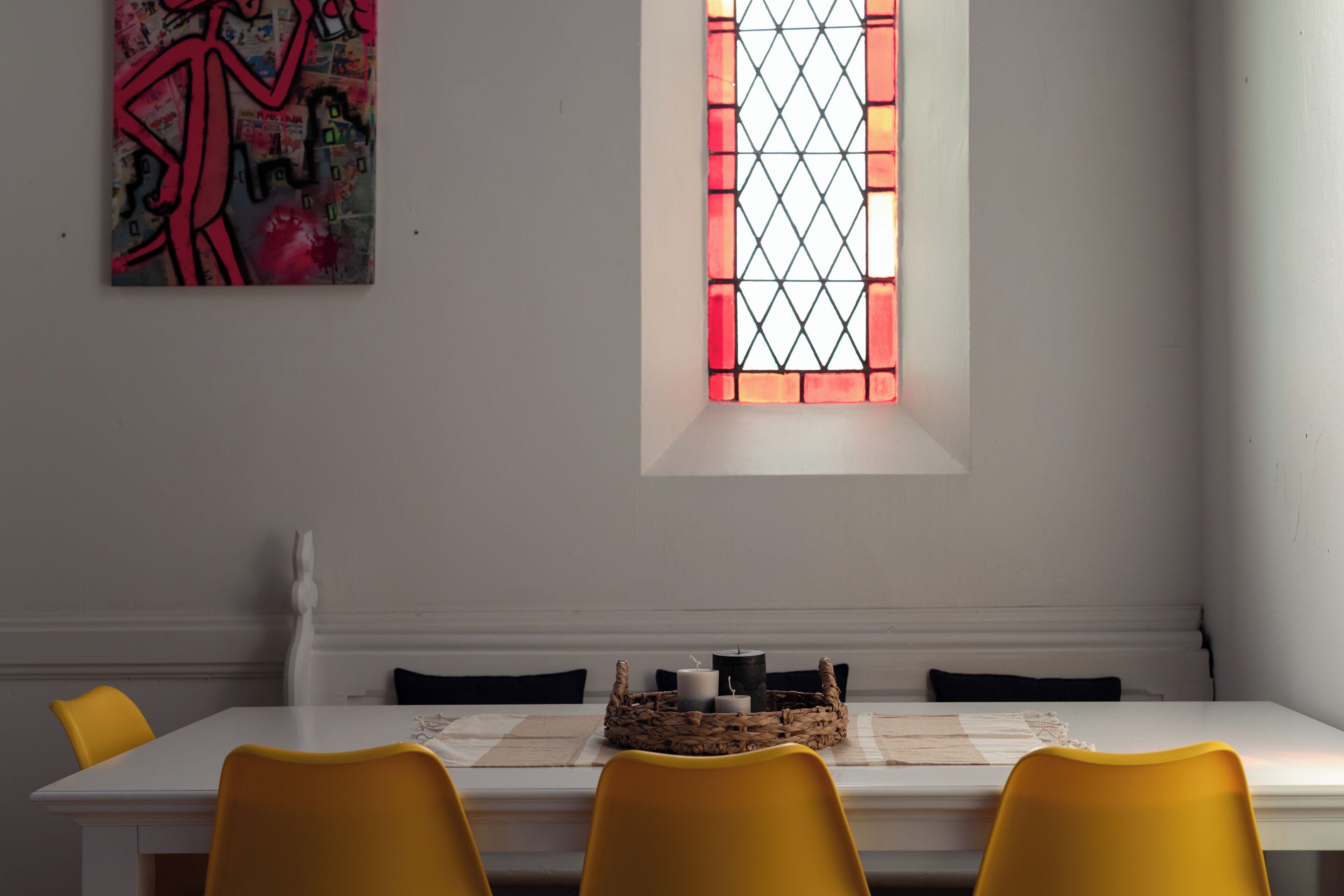 A dining area with a white table, yellow chairs, a stained glass window, and a wall-mounted artwork.