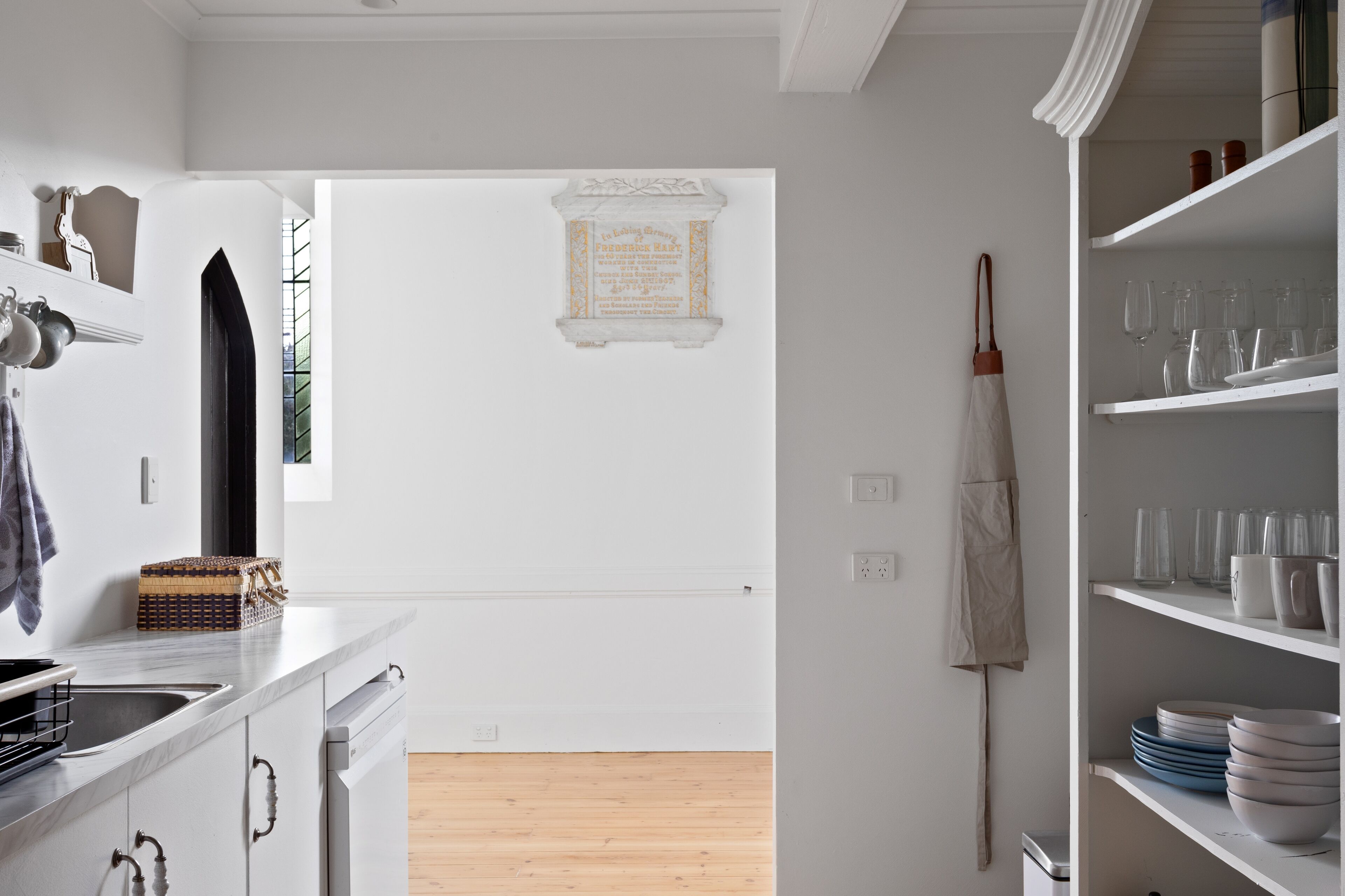 Una cocina moderna con armarios blancos, encimera de mármol y suelo de madera. Se observa un paraguas blanco colgado en la pared y una estantería con cristalería.