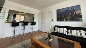 A modern living room with a glass coffee table, black sofa, and a cityscape painting. There are three black bar stools and a white door.