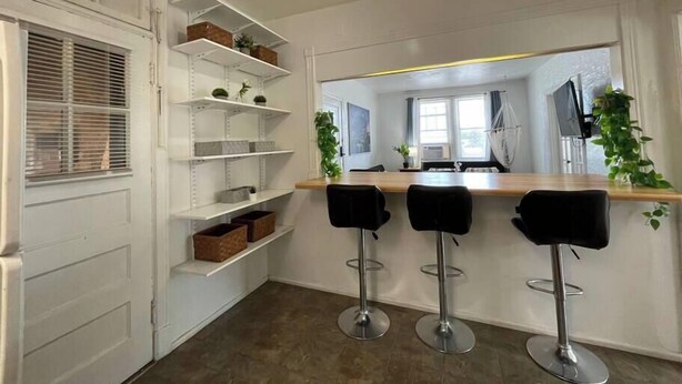 A modern kitchen with white cabinetry, a dining area with bar stools, and a view into the living room.