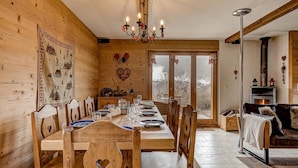 Un intérieur en bois comprenant une table à manger pour six personnes, des chaises en bois, un lustre, une cheminée et une grande fenêtre donnant sur la forêt.