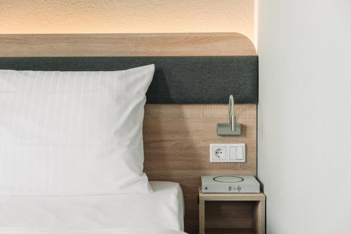 Una cama bien hecha con un almohadón blanco y un cabecero de madera con un panel oscuro. Se ve una mesita de noche con una regleta de enchufes y un pequeño dispositivo electrónico.