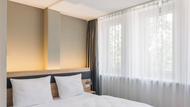 Habitación de hotel con una cama grande, cabecera de madera, mesita de noche y cortinas traslúcidas en la ventana.