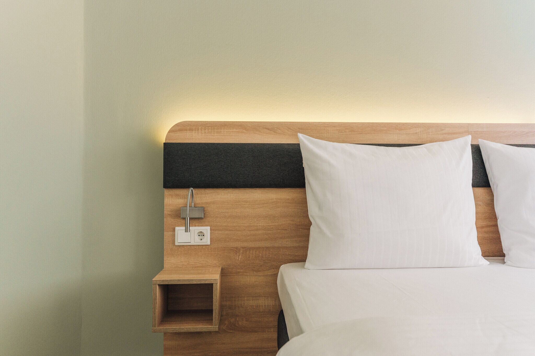 Un lit bien fait, avec du linge de lit blanc et des oreillers, une tête de lit en bois et une petite table de chevet équipée d’une prise électrique.