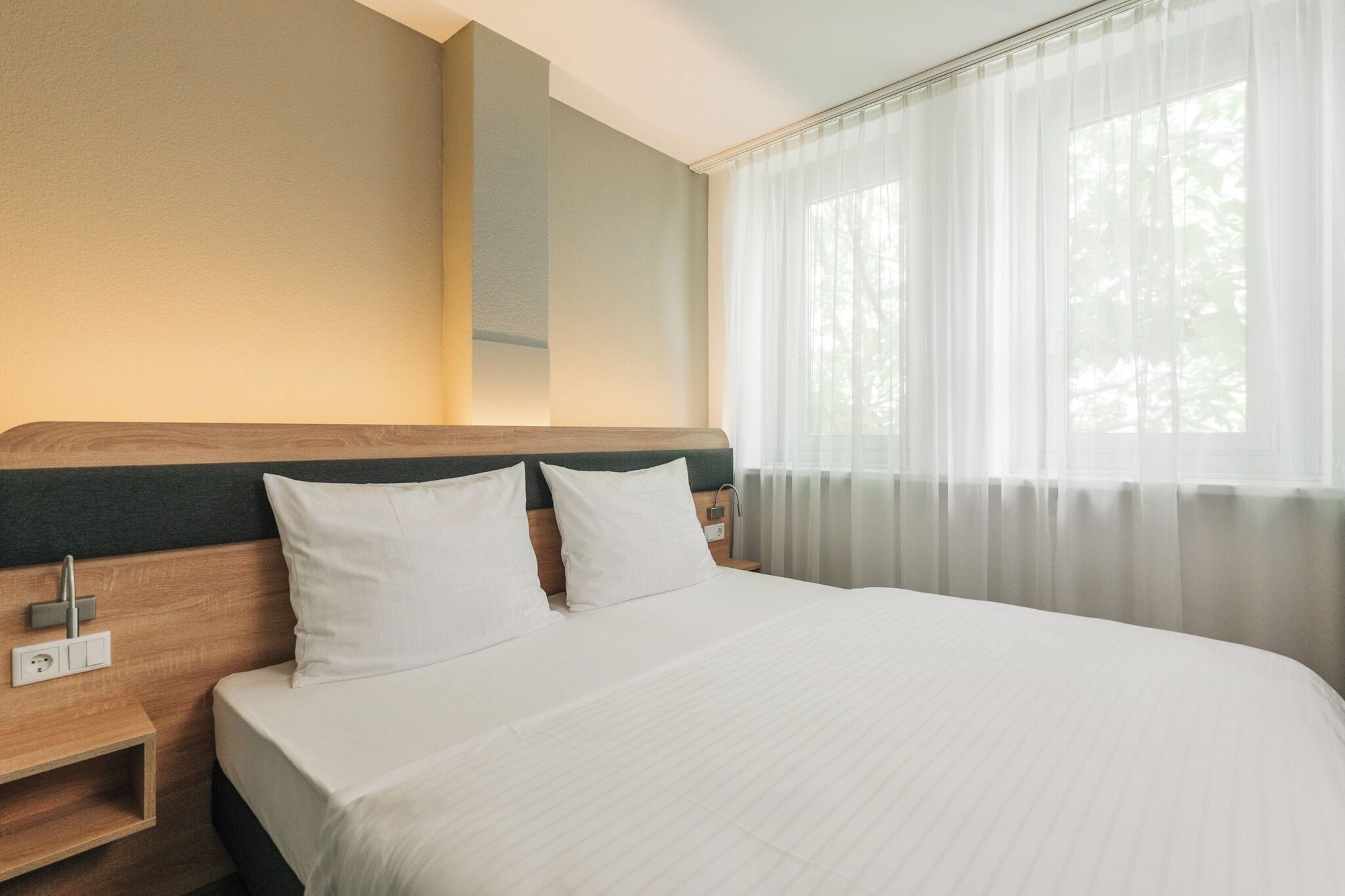 Una camera d'albergo con un letto grande, una testiera in legno, un comodino e una finestra con tende leggere.