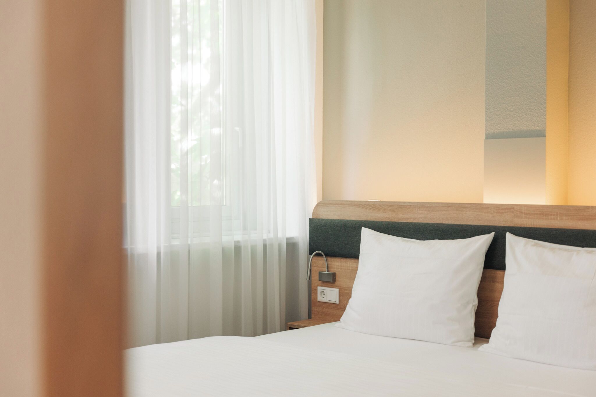 Habitación de hotel con cama, ropa de cama blanca, cabecera de madera, mesita de noche y ventana con cortinas de gasa.