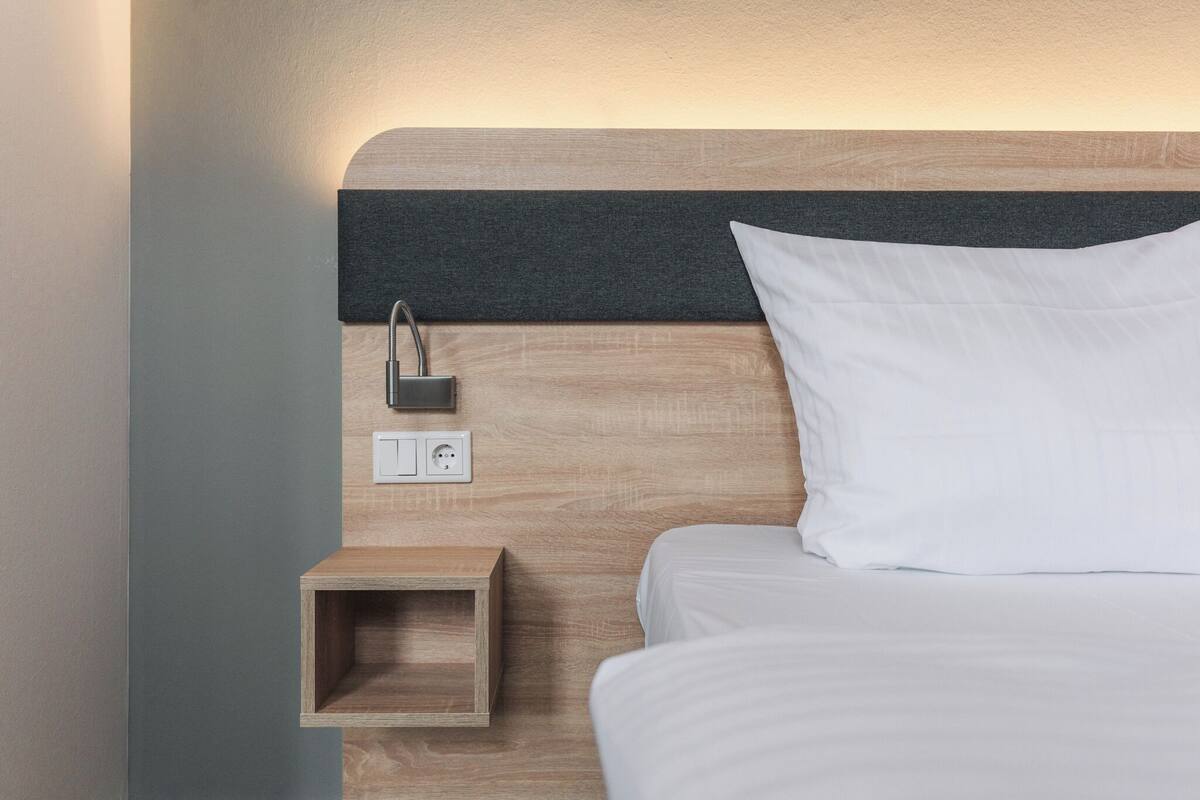A neatly made bed with white linens, a wooden nightstand with a power outlet, and a wall-mounted lamp.