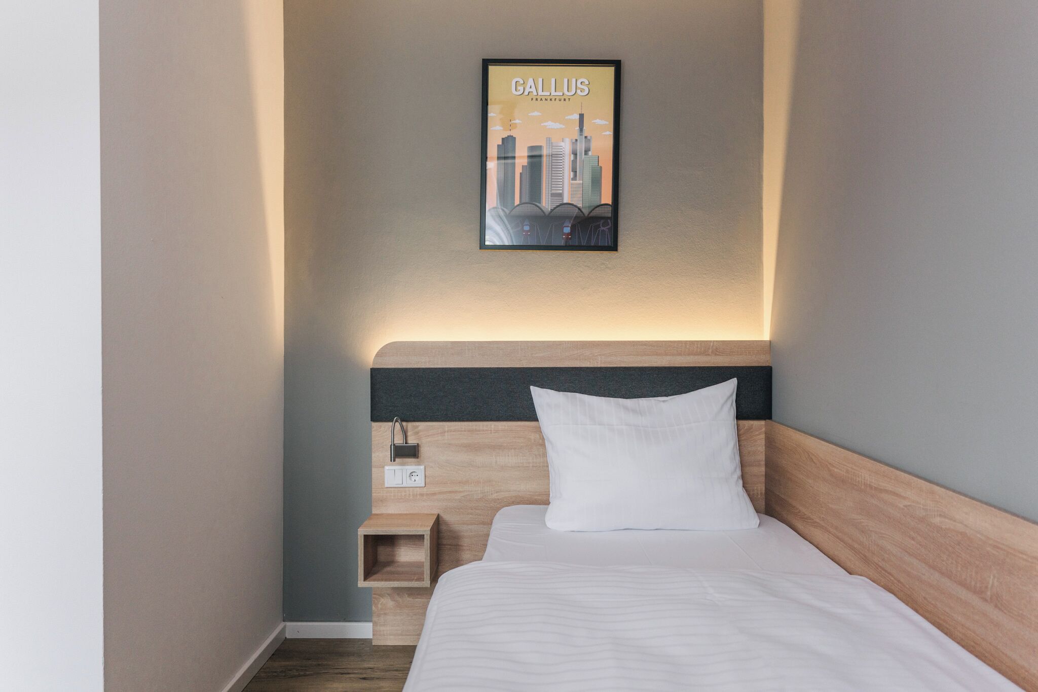 Una habitación de hotel con una cama, una mesita de noche, un cartel con un paisaje urbano enmarcado y un enchufe en la pared.