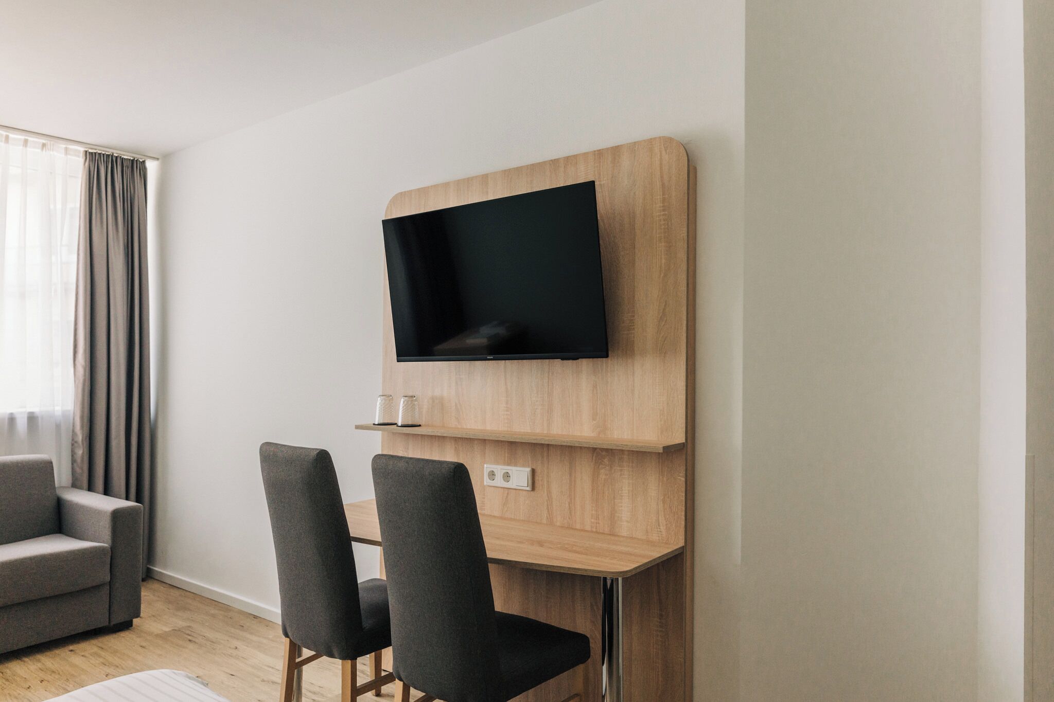 A modern hotel room with a mounted TV, a grey sofa, two chairs, and a wooden desk.