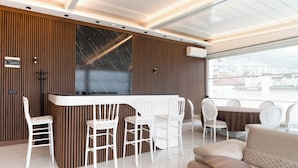 Un interior moderno con una zona de bar, sillas blancas, un sofá y vistas a la ciudad a través de amplias ventanas.