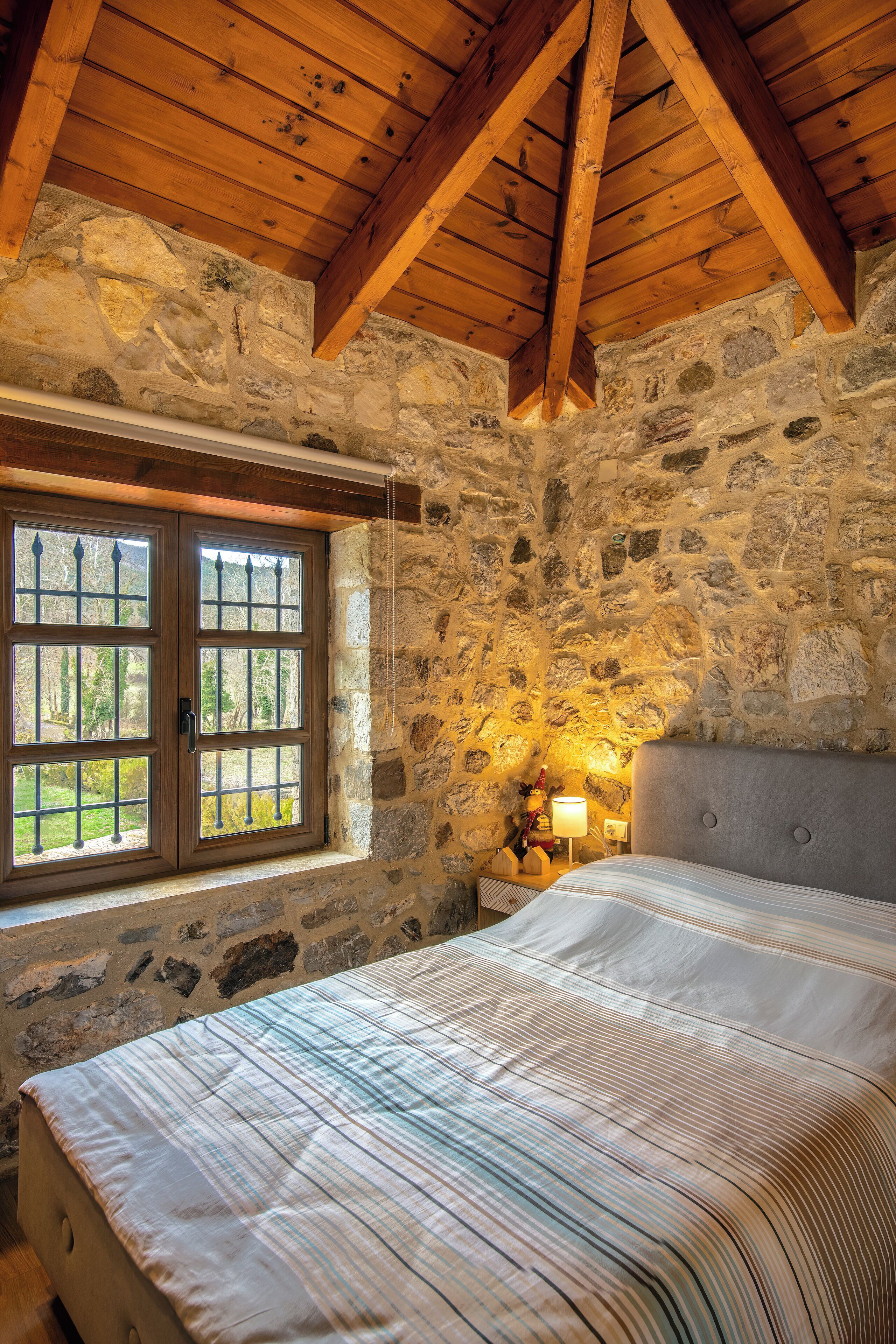 A bedroom with a stone wall, wooden beams, a bed with a striped bedspread, and a window with a view of greenery.