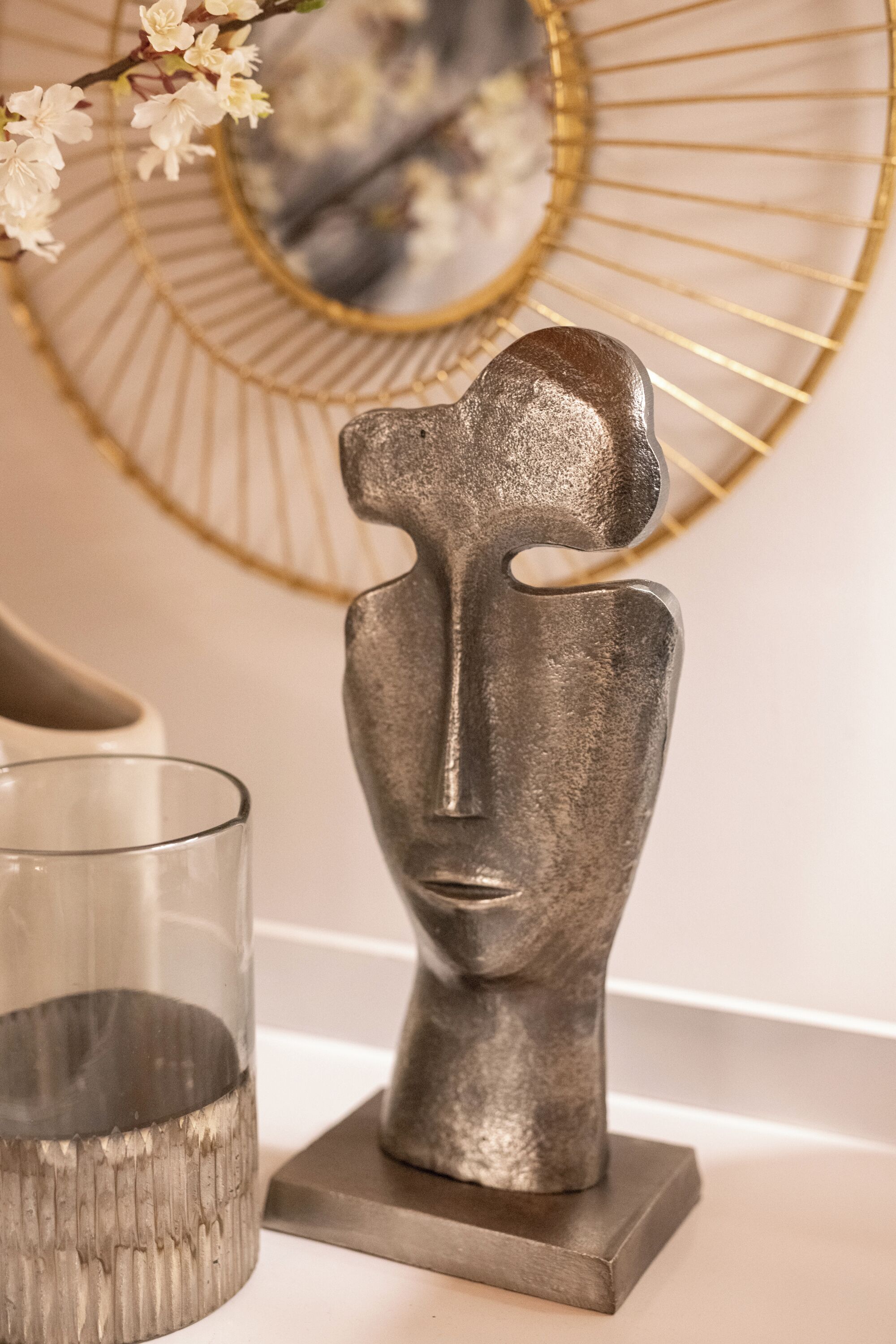 Una escultura metálica de un rostro humano sobre una base rectangular, un recipiente de vidrio y un espejo con marco dorado sobre una superficie blanca.