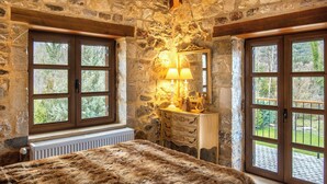 Un dormitorio con pared de piedra, vigas de madera en el techo, una cama grande con cubrecama de piel, una mesita de noche, una lámpara y una ventana con vistas a árboles.