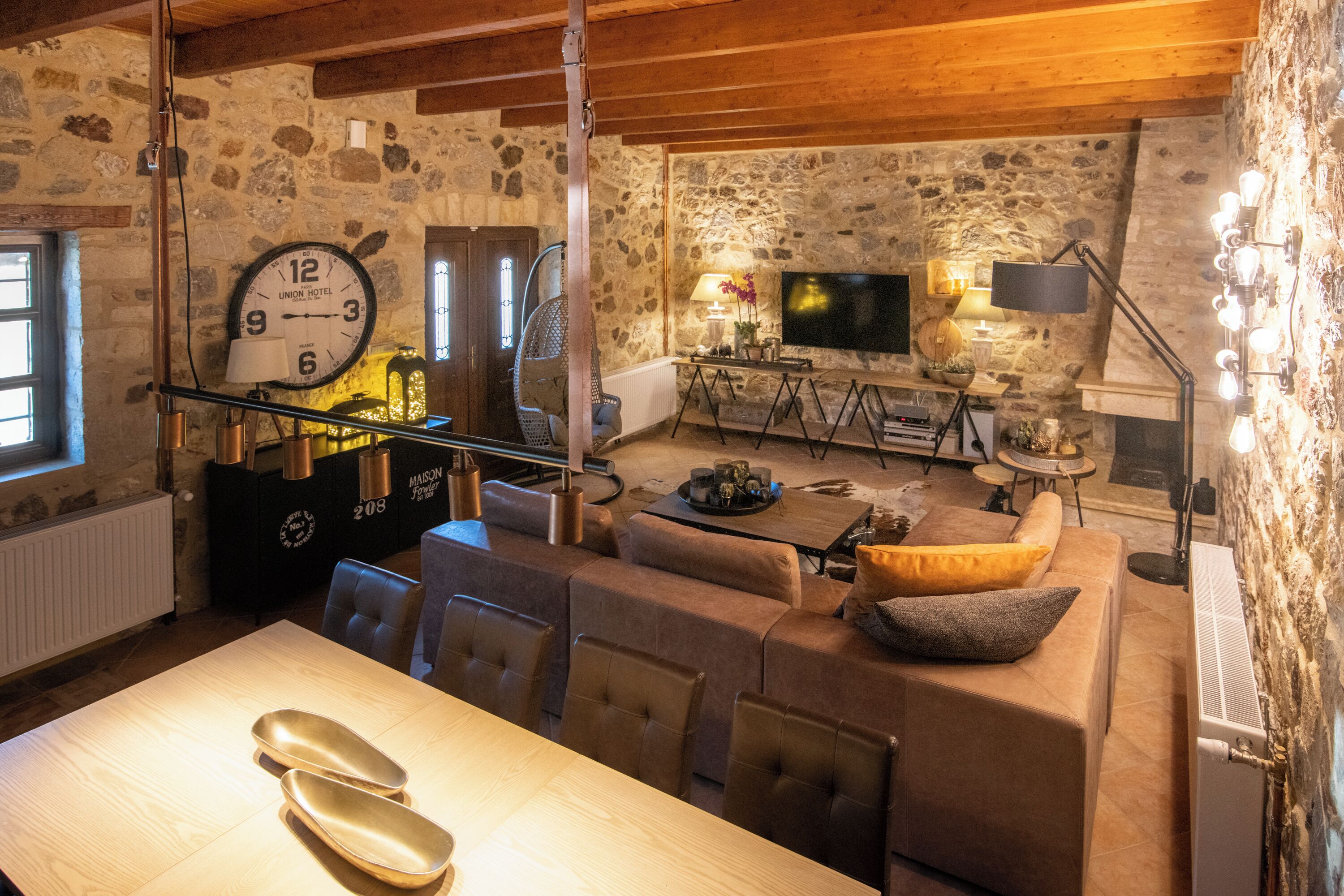 Una sala de estar acogedora con pared de piedra, vigas de madera en el techo, un reloj grande, un televisor de pantalla plana y un comedor con mesa y sillas.