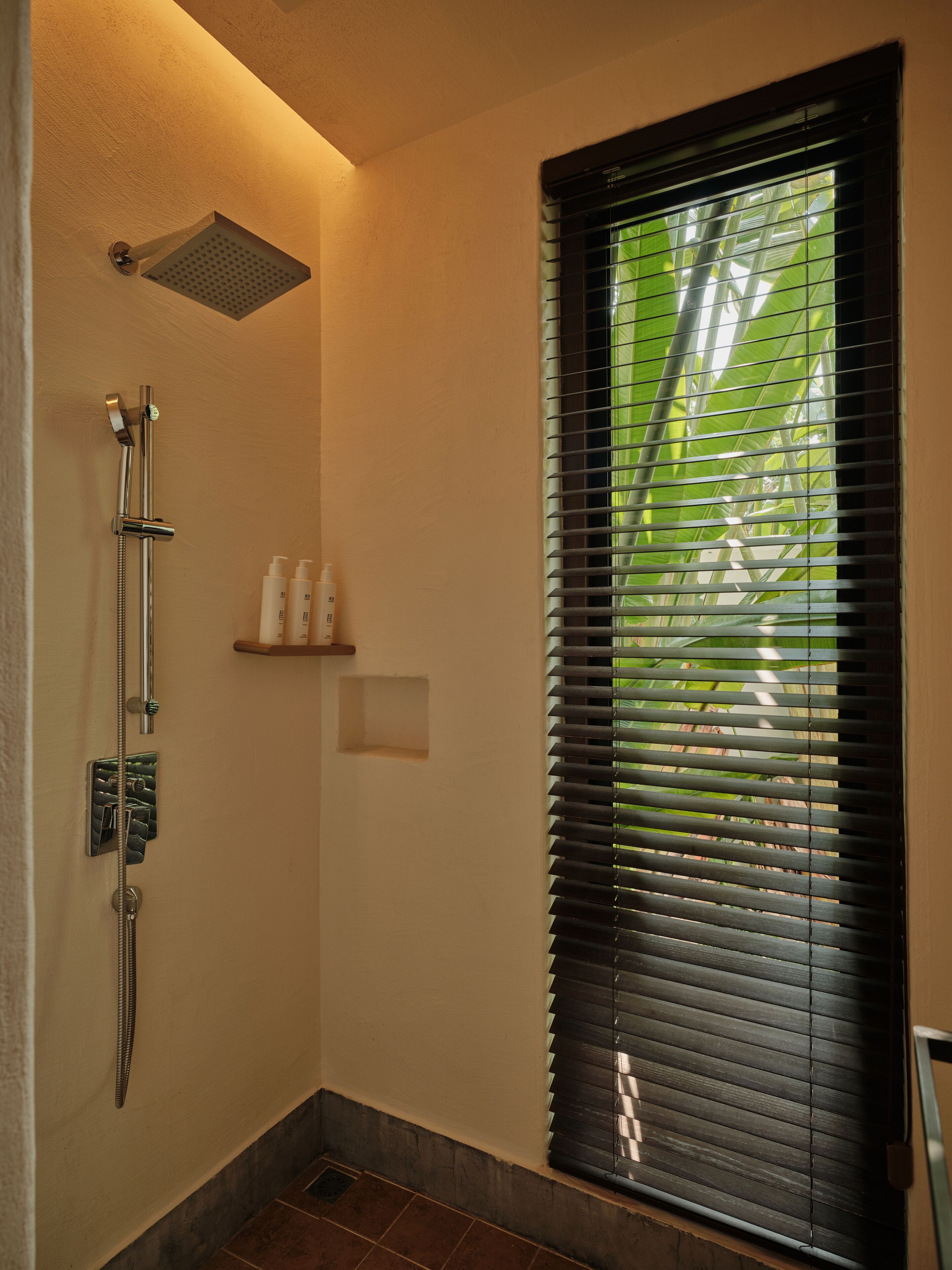 A bathroom with a shower, a window with blinds, and a shelf with toiletries.