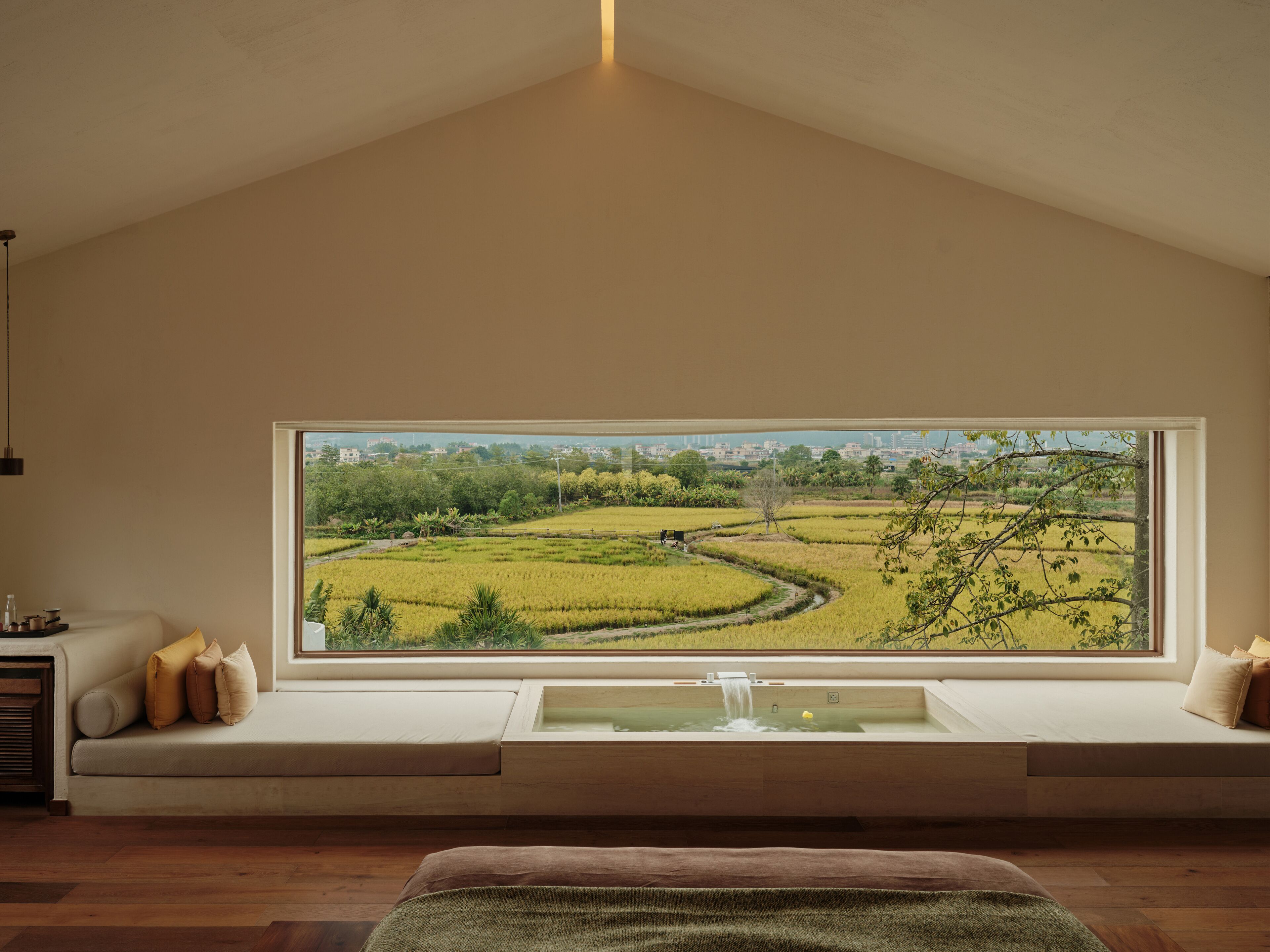 Una habitación con un ventanal grande con vistas a un campo de arroz, un banco empotrado con cojines y una pequeña fuente.