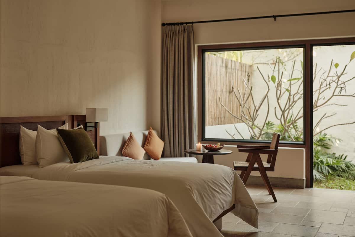 A hotel room with two beds, a small table, a chair, and a large window with a view of greenery.
