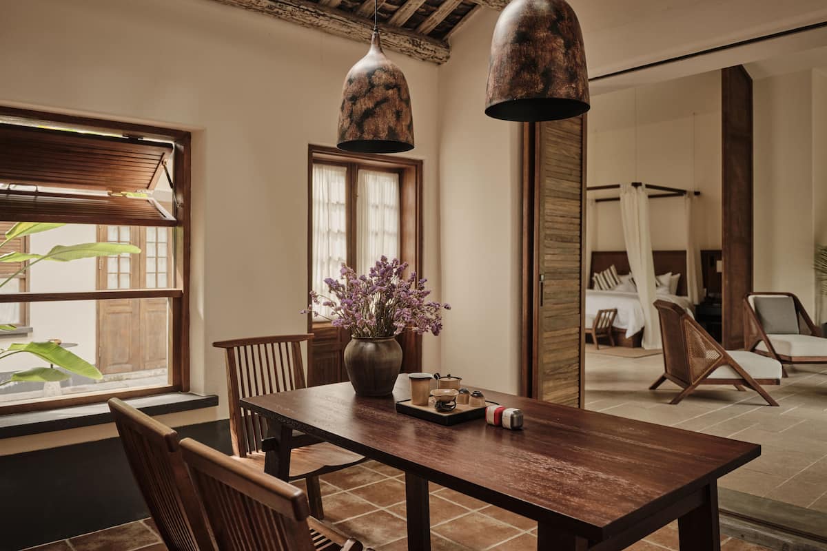 A wooden dining table with chairs, a vase of flowers, and hanging pendant lights in a room with wooden beams and a view of a bedroom with a bed and curtains.