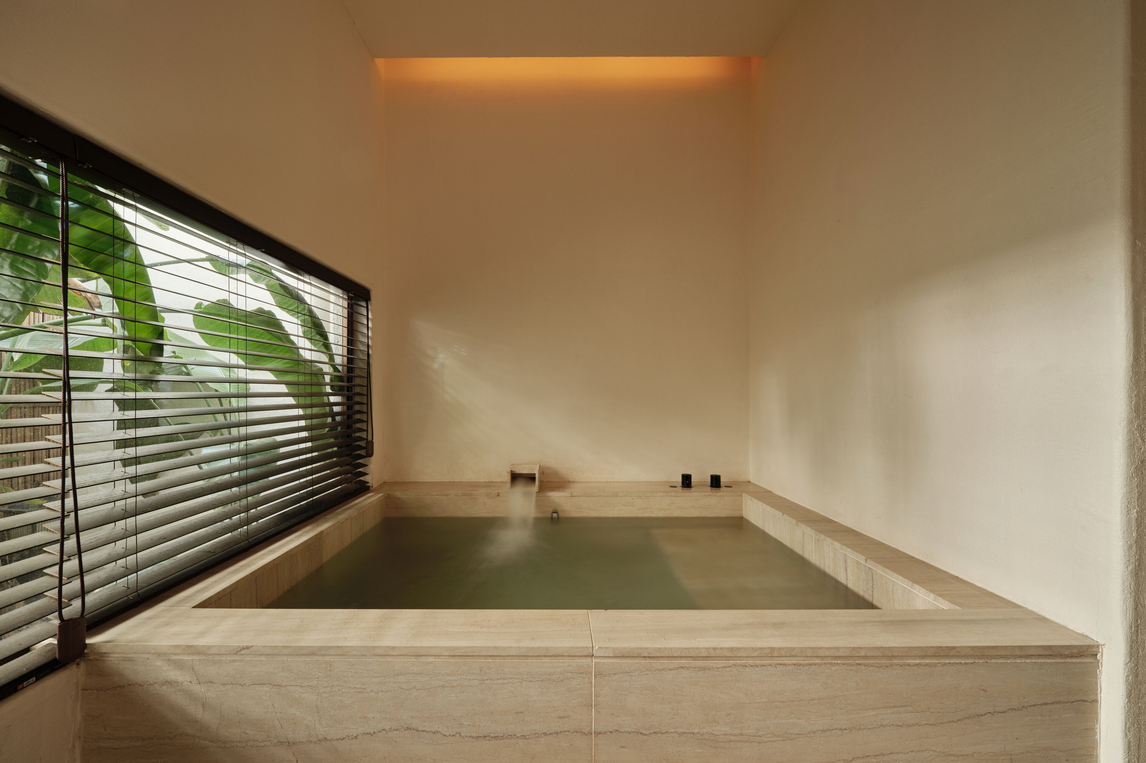 A modern spa room with a rectangular hot tub, a window with blinds, and a small sink.