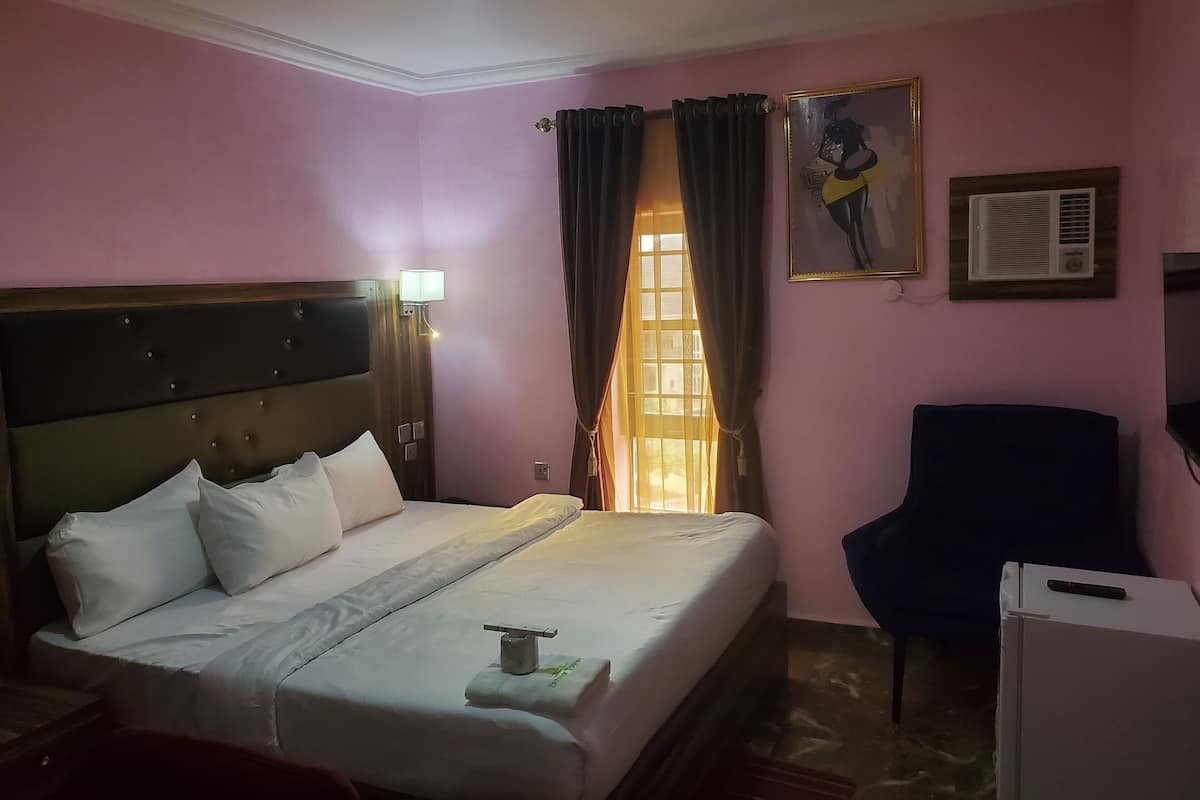 Una habitación de hotel con cama, una silla, un cuadro, una ventana con cortinas y un equipo de aire acondicionado.