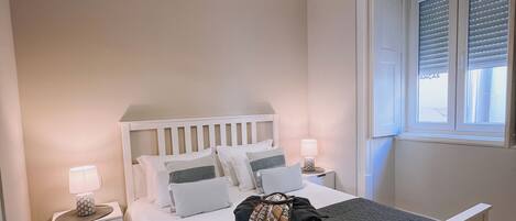 Un dormitorio con cama, mesitas de noche, lámpara, ventana con persianas y una lámpara colgante.