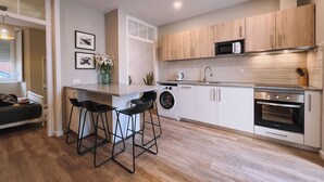 A modern kitchen with a built-in oven, microwave, and washing machine. There are bar stools and a small seating area with a window.
