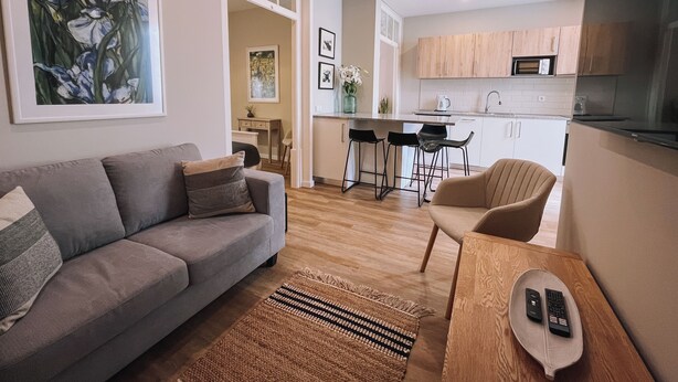 A modern living room with a grey sofa, a wooden coffee table, and a kitchen area with wooden cabinets and a microwave.