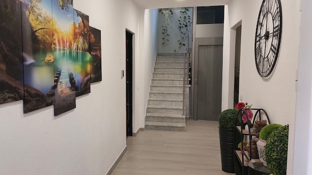 Un couloir avec un escalier, une horloge murale et une composition florale dans un pot.